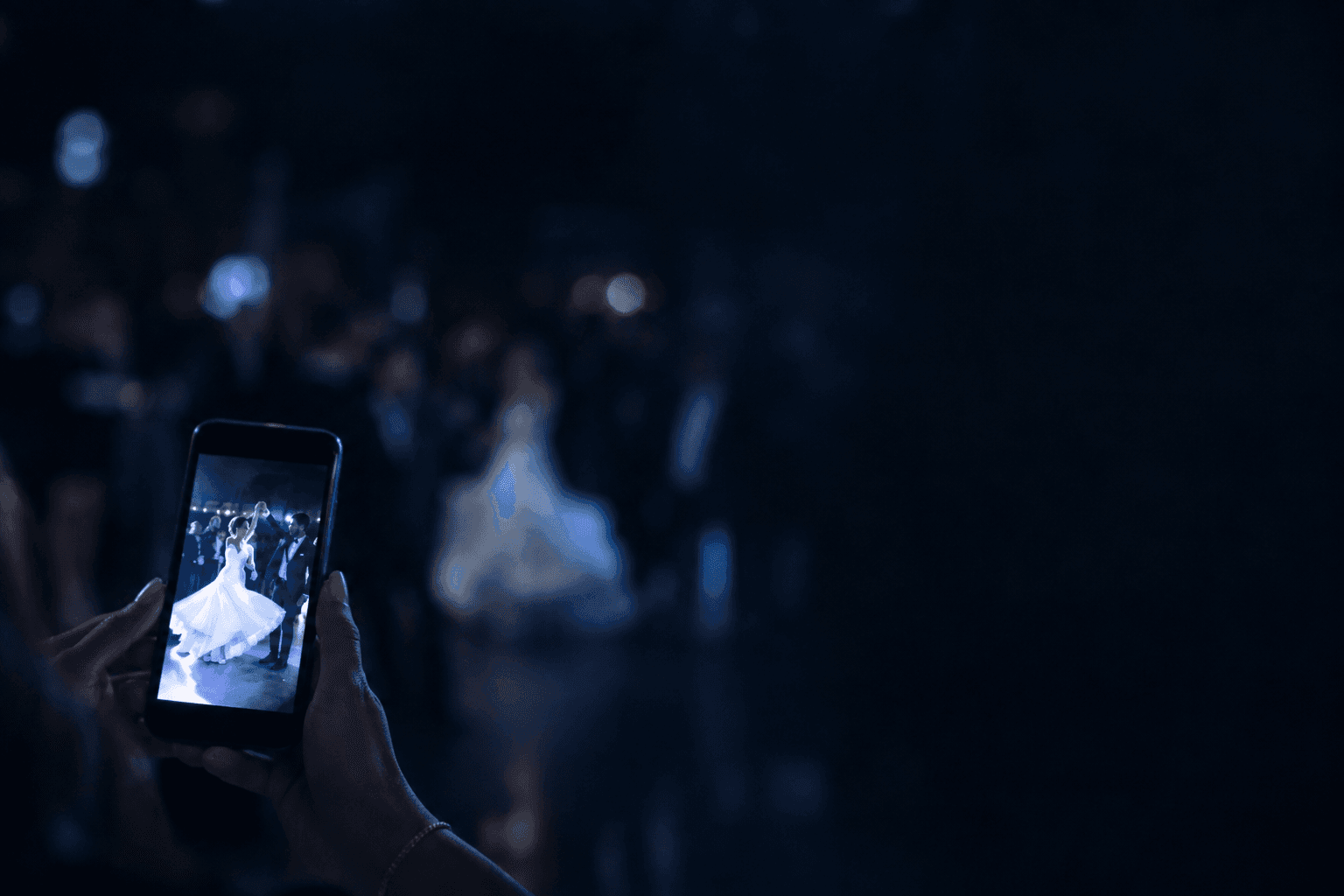 Un'invitata fotografa con lo smartphone gli sposi che ballano il primo ballo, il telefono illuminato in basso a sinistra su una pista buia