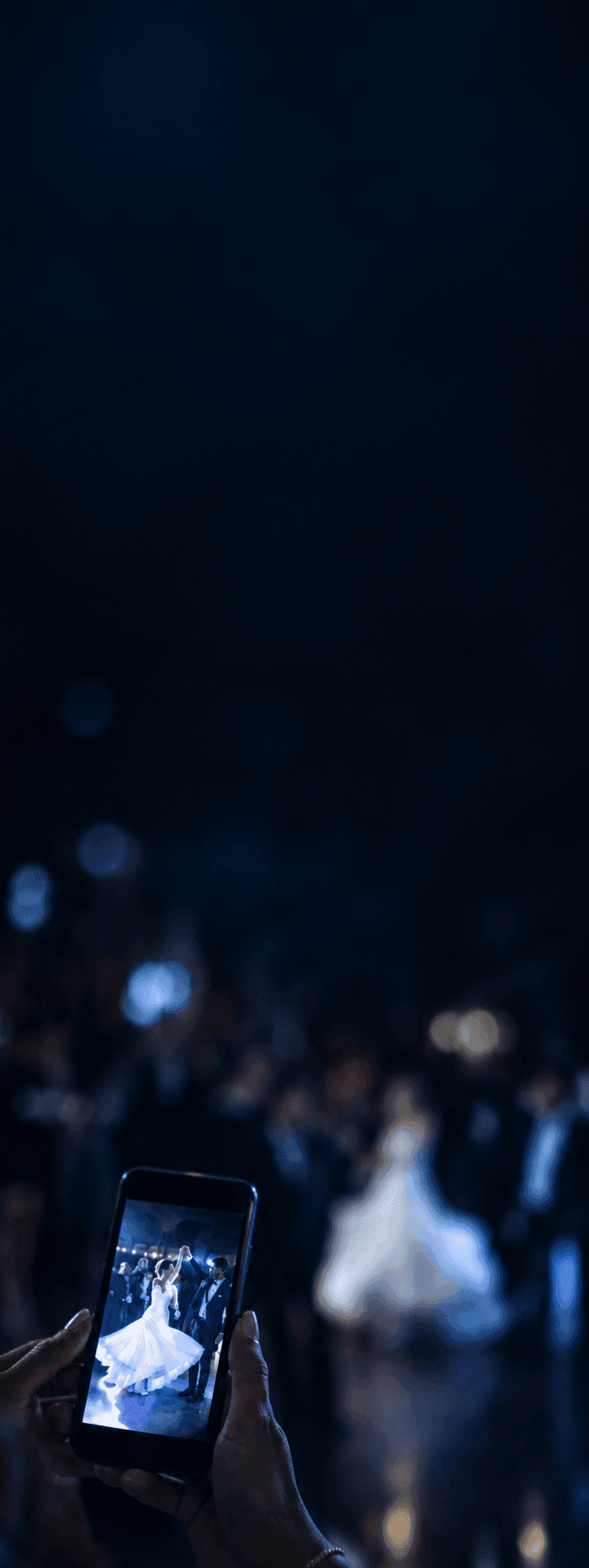 Un'invitata fotografa con lo smartphone gli sposi che ballano il primo ballo, il telefono illuminato in basso a sinistra su una pista buia