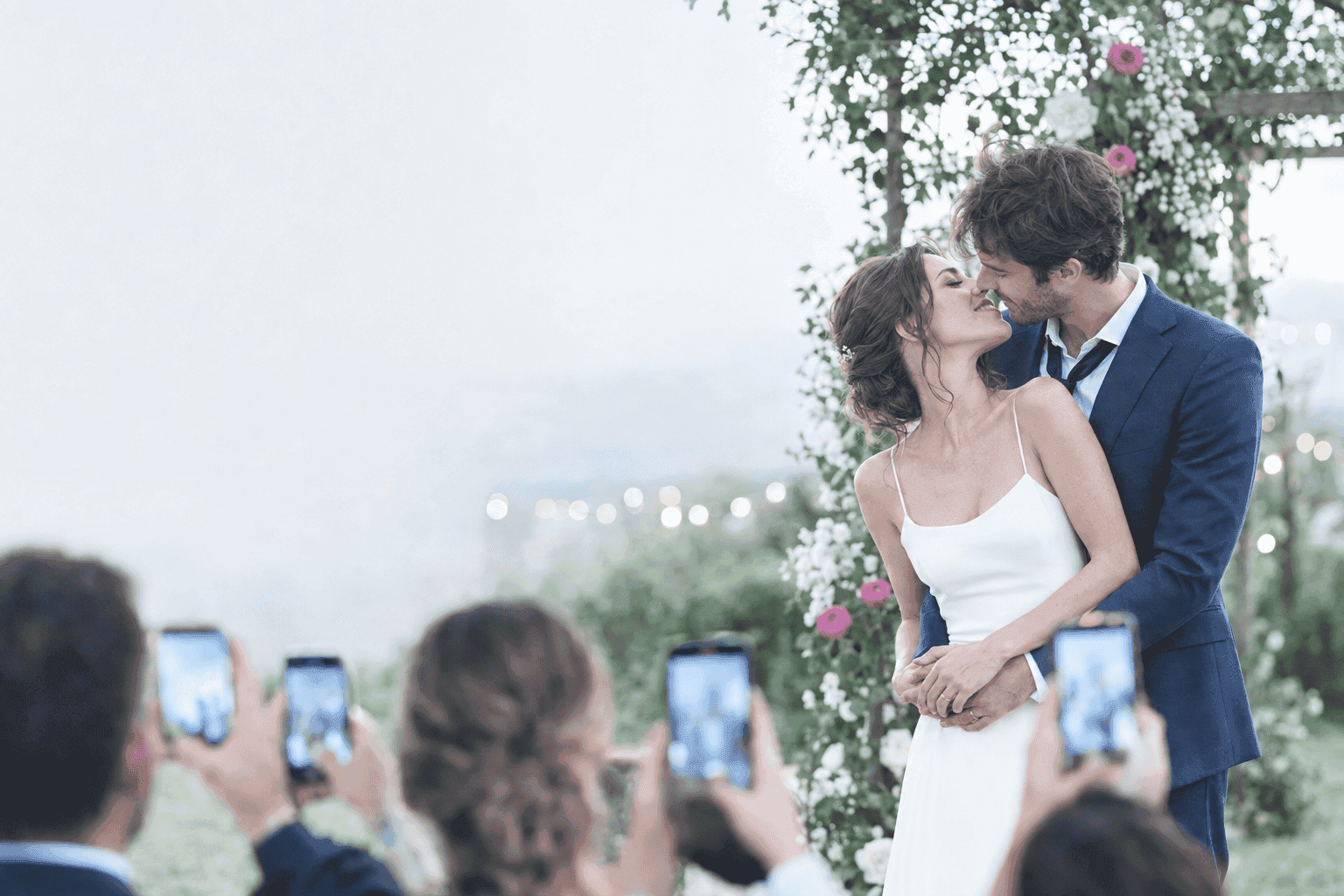 Sposi che si baciano durante la cerimonia, circondati da invitati che fotografano il momento con lo smartphone